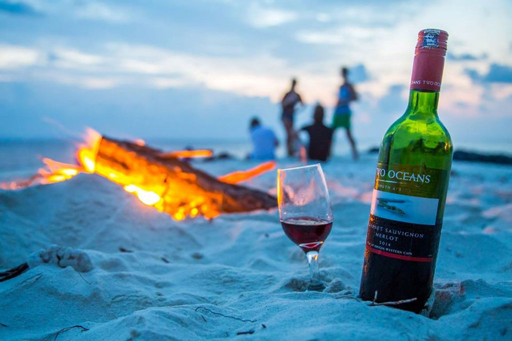 Bottle of wine and glass on a beach witha bonfire and party goers in the background